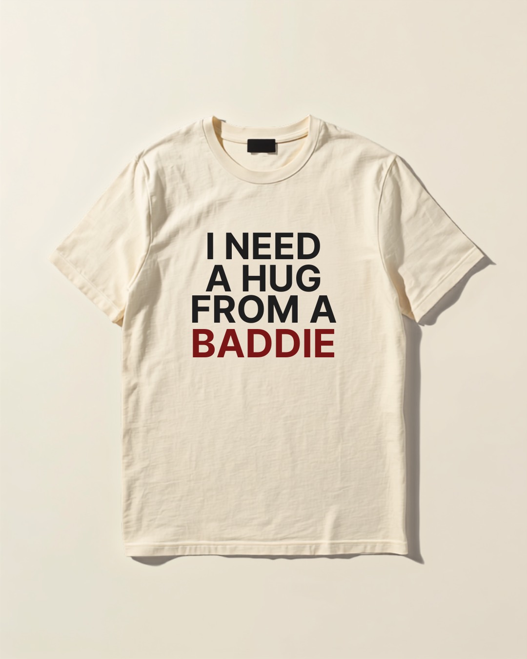 Ivory heavyweight tee reading I NEED A HUG FROM A BADDIE, flat lay studio shot